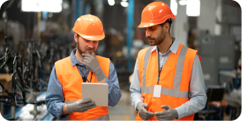 Two engineers in safety gear reviewing plans on a tablet in a factory